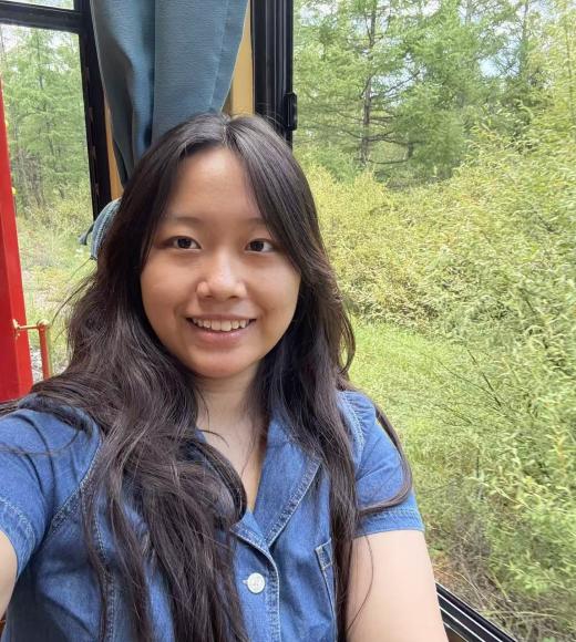 Young woman with long dark hair smiling inside a train, green trees visible outside.